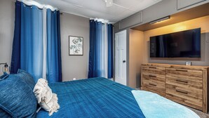 A bedroom with a bed, a wooden dresser, a TV, and blue curtains. - Jackson Street Suites #1 (Dubuque)