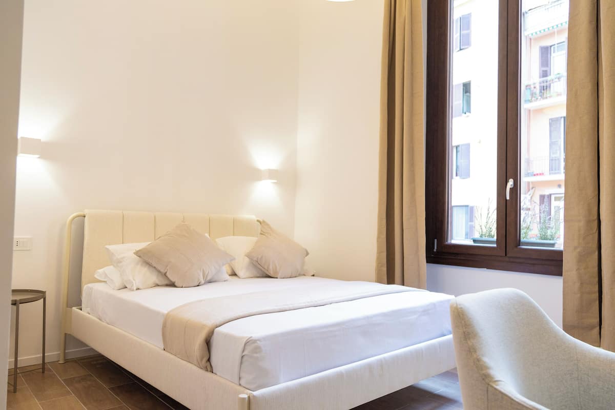 A neatly made bed with a headboard, a bedside table, and a chair in a room with a window and curtains.
