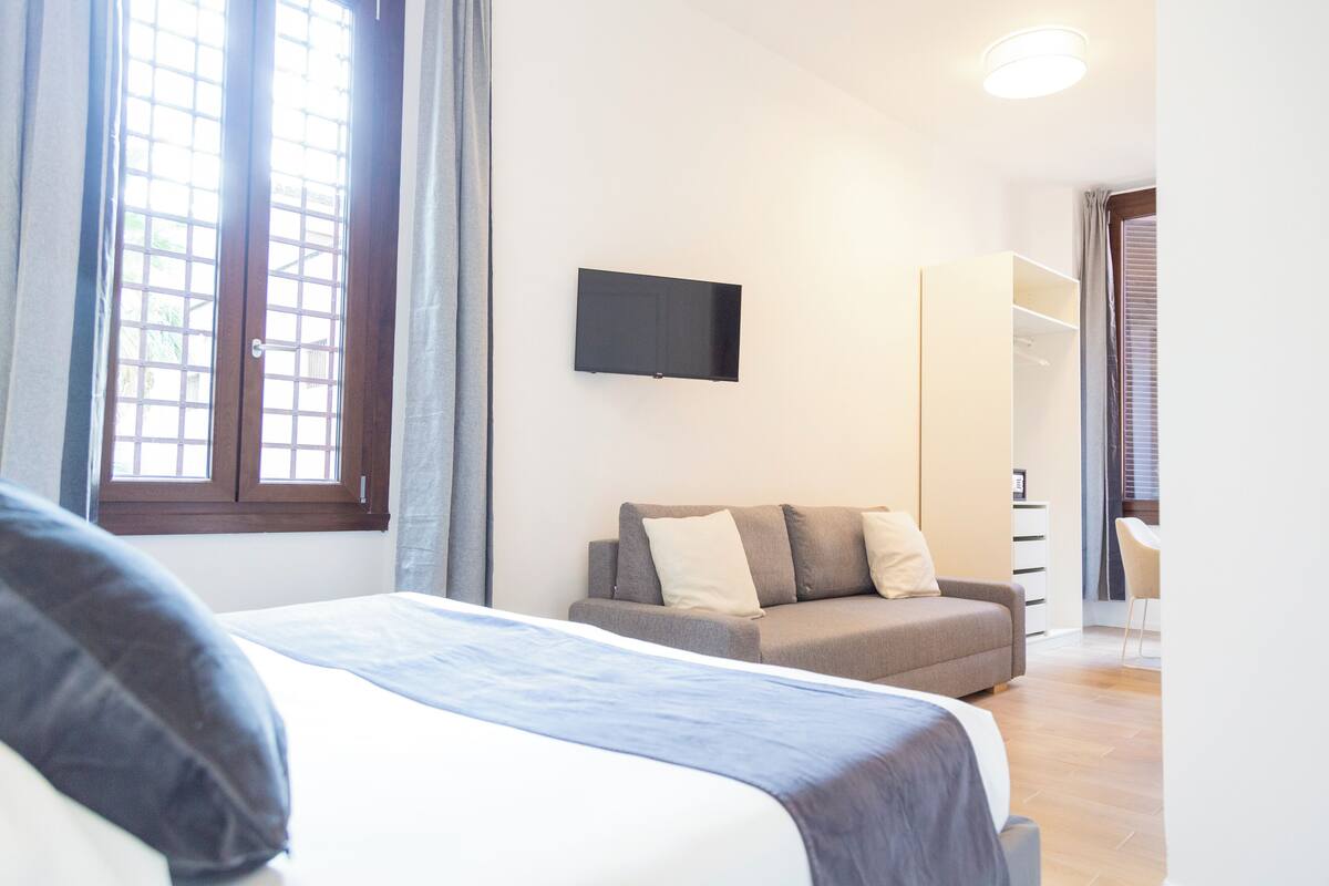 A bedroom with a bed, a sofa, a TV mounted on the wall, and a wardrobe.
