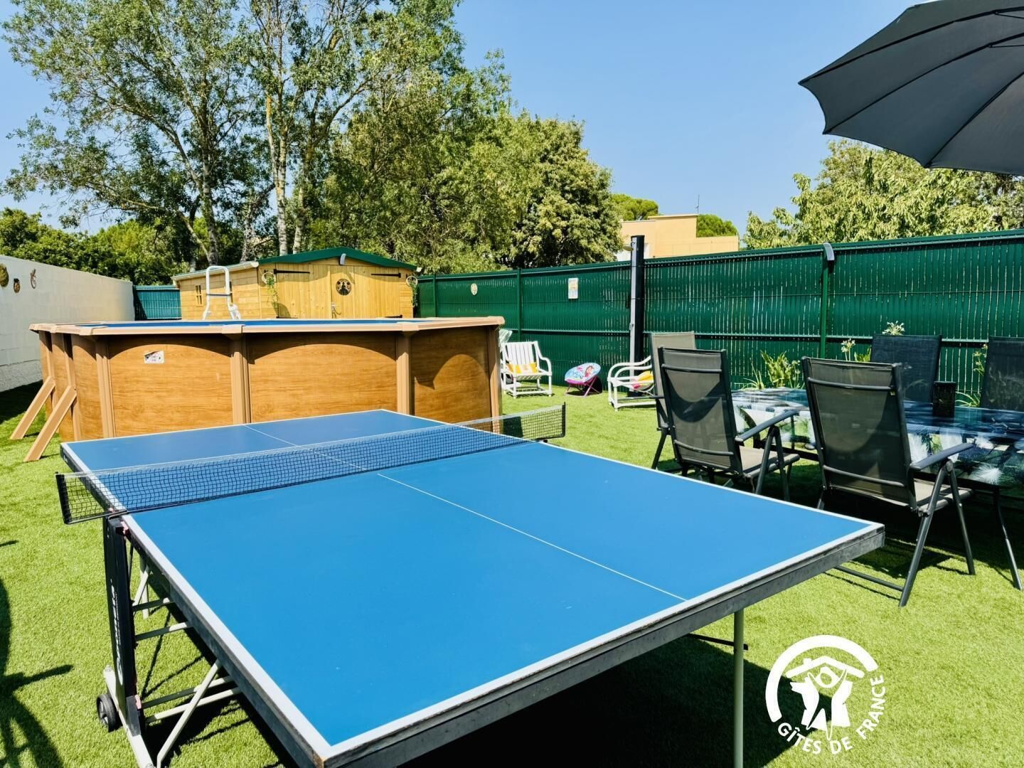 An outdoor ping pong table set up in a backyard with a green fence, chairs, and a small wooden structure.