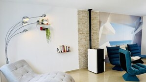 A modern living room with a curved sofa, a floor lamp, a bookshelf with books and plants, and a large window with a view of the sea.