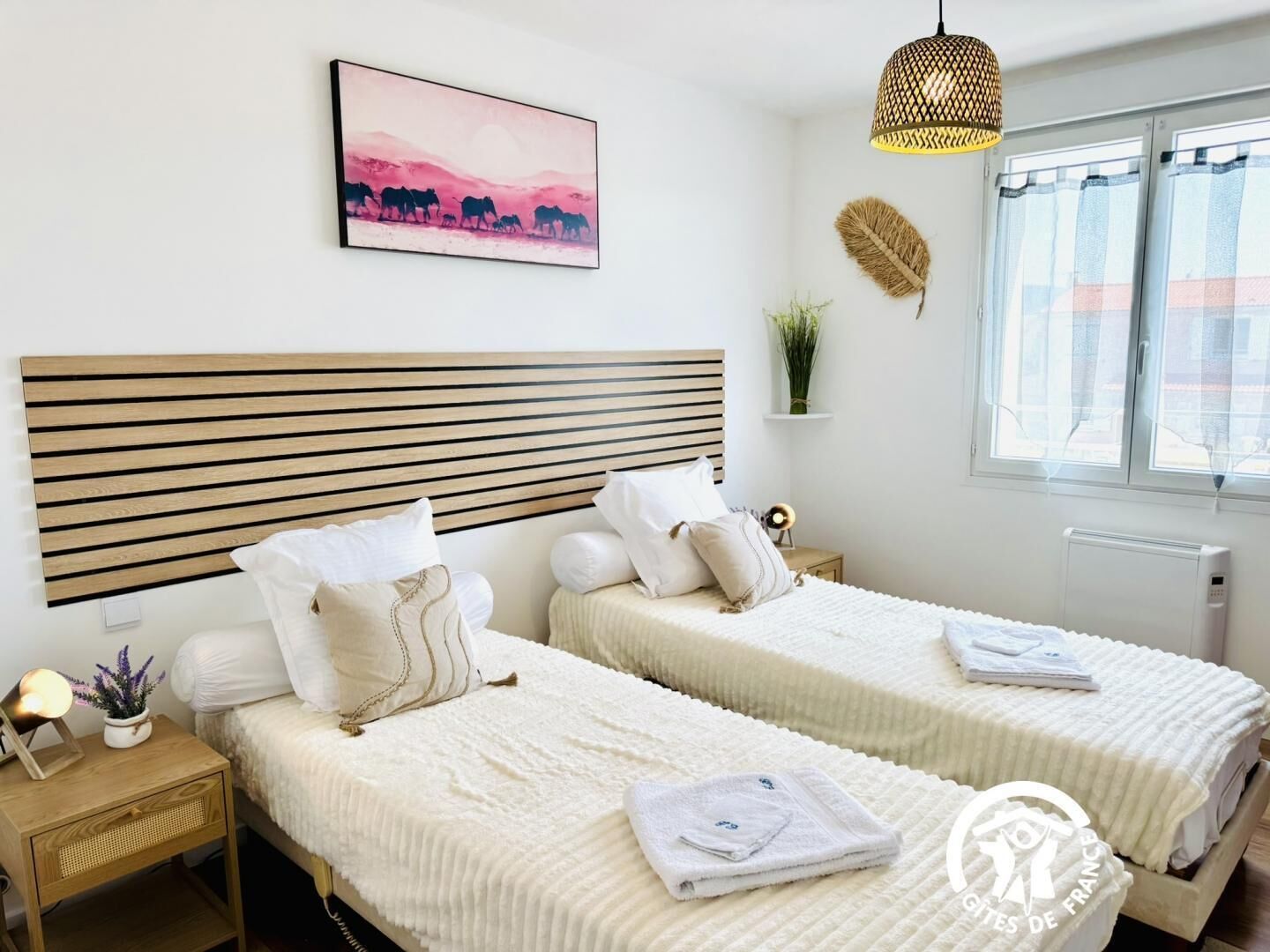 A hotel room with two beds, a wooden headboard, a bedside table with a lamp, a wall-mounted picture, a window with curtains, and a radiator.