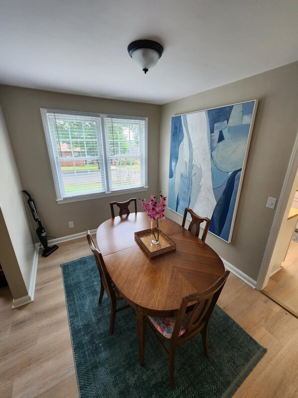 A dining area with a round wooden table, six chairs, a vase with pink flowers, and a large abstract painting on the wall.