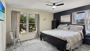 Ein Schlafzimmer mit Bett, einem Sessel, einem Deckenventilator und einem Fenster mit Vorhängen.