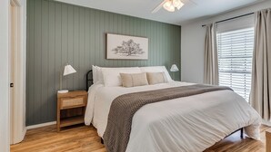 Une chambre à coucher comprenant un lit, des tables de chevet, un ventilateur de plafond, un tableau encadré et une fenêtre avec des rideaux.