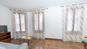 Una habitación con una cama, una mesa de madera, un televisor sobre un mueble y cortinas con un estampado ondulado.