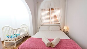 Un dormitorio con una cama blanca, una colcha rosa y una cuna blanca llena de juguetes.