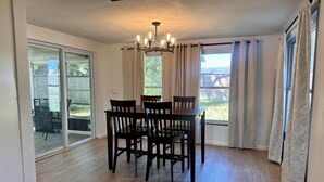 A dining area with a wooden table and chairs, a chandelier, and large windows with curtains. - Sunny 2BR Retreat · Enclosed Patio · Pet Friendly · Near Gulf Beaches (Port Richey)