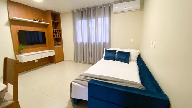 Un dormitorio con una cama, un televisor instalado en un mueble de madera, una ventana con cortinas y un equipo de aire acondicionado.