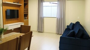 Una sala de estar con televisor montado en la pared, una mesa de comedor con sillas, un sofá azul y una ventana con cortinas.