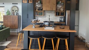 A modern kitchen with dark cabinetry, a central island with stools, and a dining area with a green sofa. - Stunning views, Luxurious, Peaceful, dog friendly, king beds,fire pit,WIFI,AC,EV (Townshend)