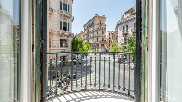 A view from a balcony overlooking a city street with historic buildings, a roundabout, and a clear sky.