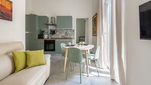 A modern living room with a sofa, a dining table with chairs, a kitchen area, and a large window with curtains.