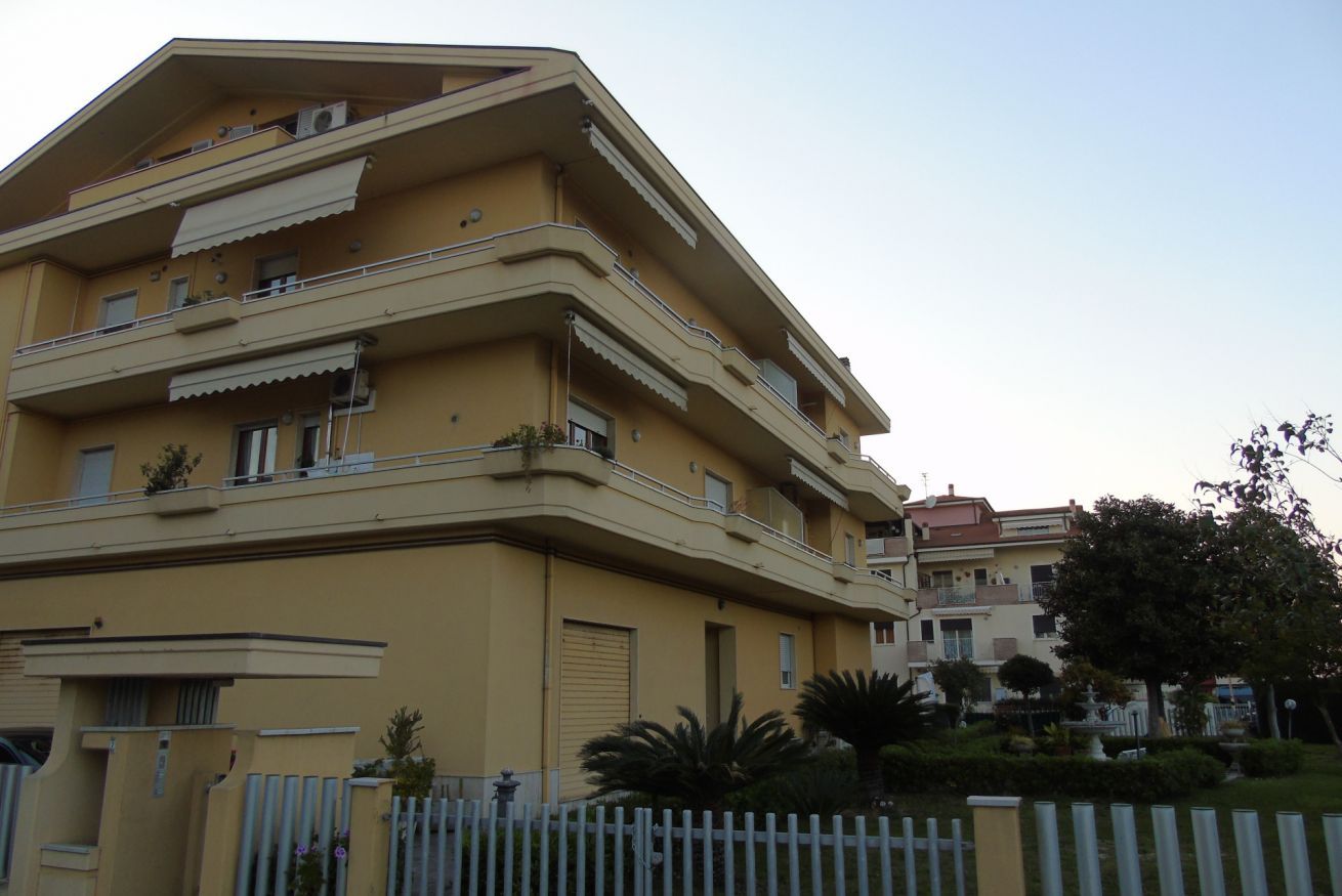 A multi-story building with balconies, a gated entrance, and a clear sky.