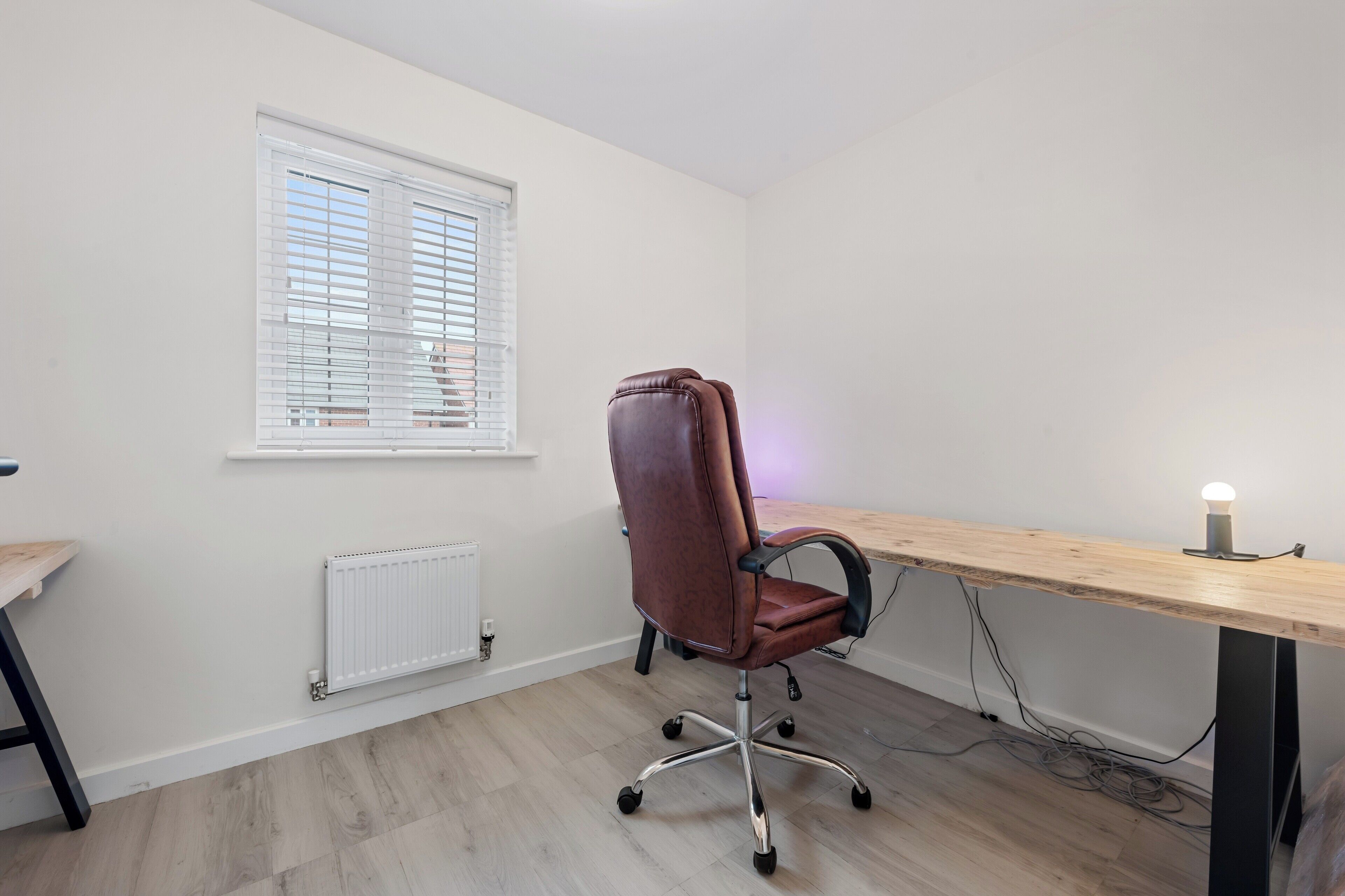 Une pièce avec un bureau en bois, un fauteuil de bureau marron, une fenêtre avec des stores et un radiateur.