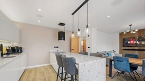 Une cuisine moderne dotée d’un îlot central, de tabourets de bar, d’un coin repas avec une table en bois et des chaises bleues, ainsi qu’un téléviseur fixé au mur affichant le logo de Netflix.