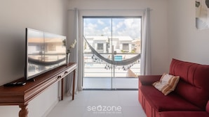 A living room with a TV on a wooden stand, a red sofa, a hammock, and a sliding glass door leading to a balcony with a view of buildings.