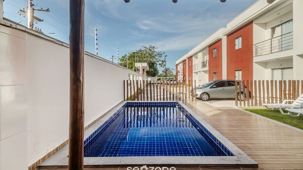 A swimming pool with a wooden deck, a car parked nearby, and a building with balconies in the background.