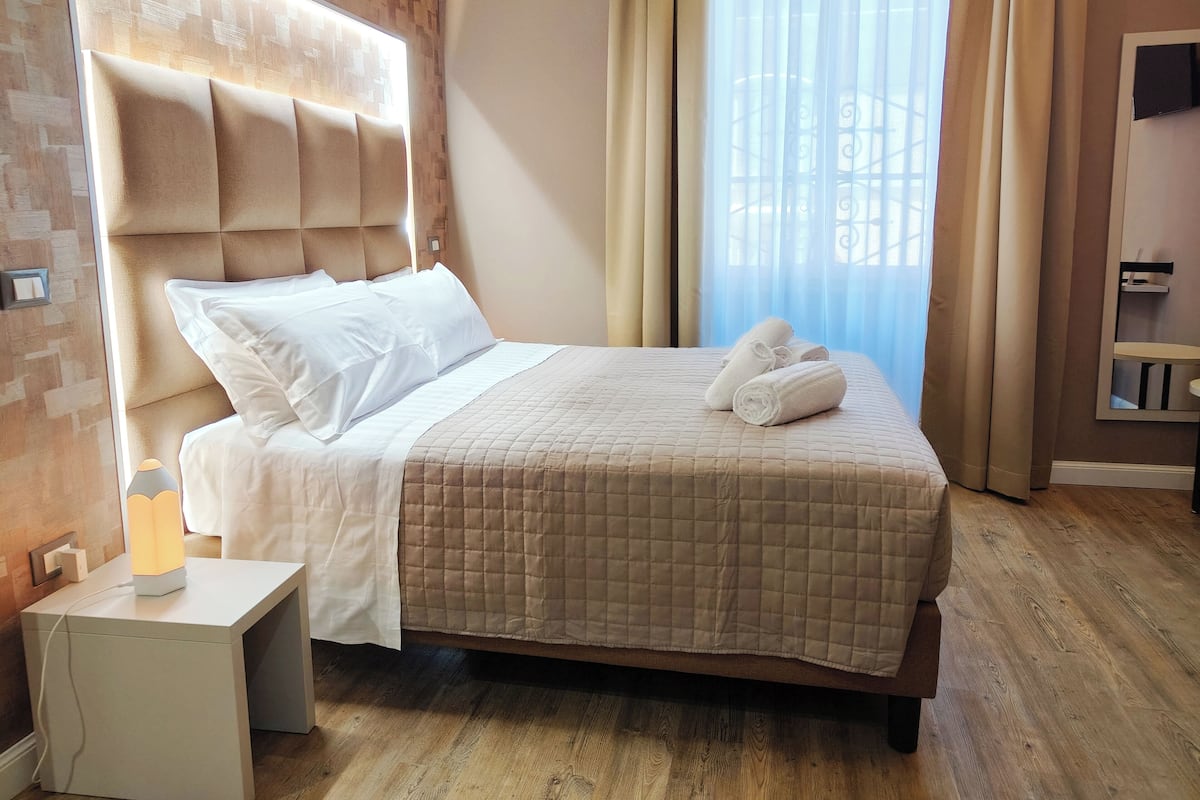 A neatly made bed with a quilted bedspread and white pillows, a bedside table with a lamp, and a window with sheer curtains.
