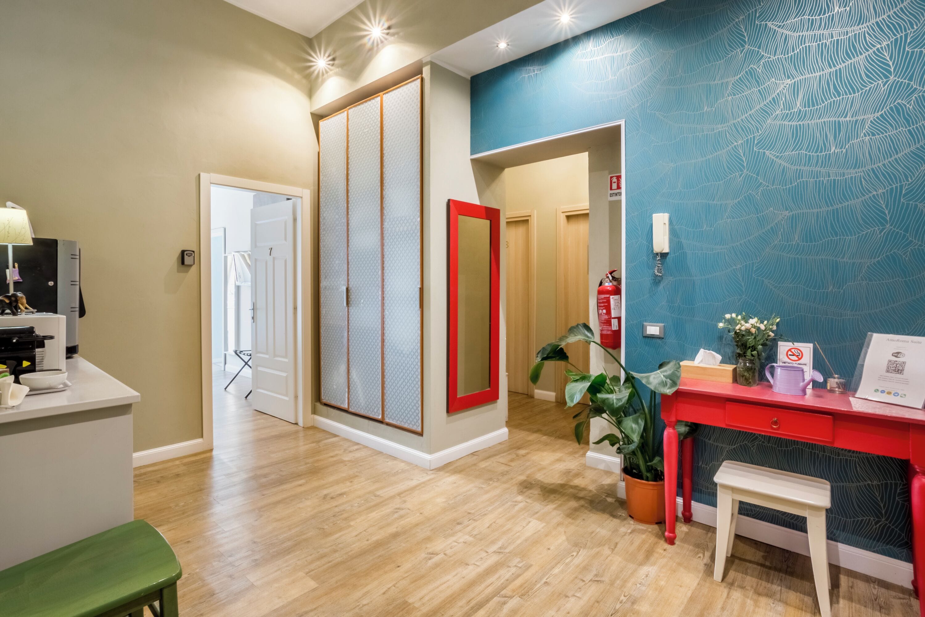 Une chambre d’hôtel avec un plancher en bois, un bureau rouge, un miroir, une plante et un mur bleu à la texture particulière.