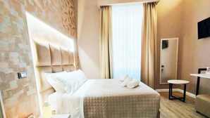 A hotel room with a bed, bedside table, a small round table, a mirror, and a window with curtains. - Nicole Suite (Rome)