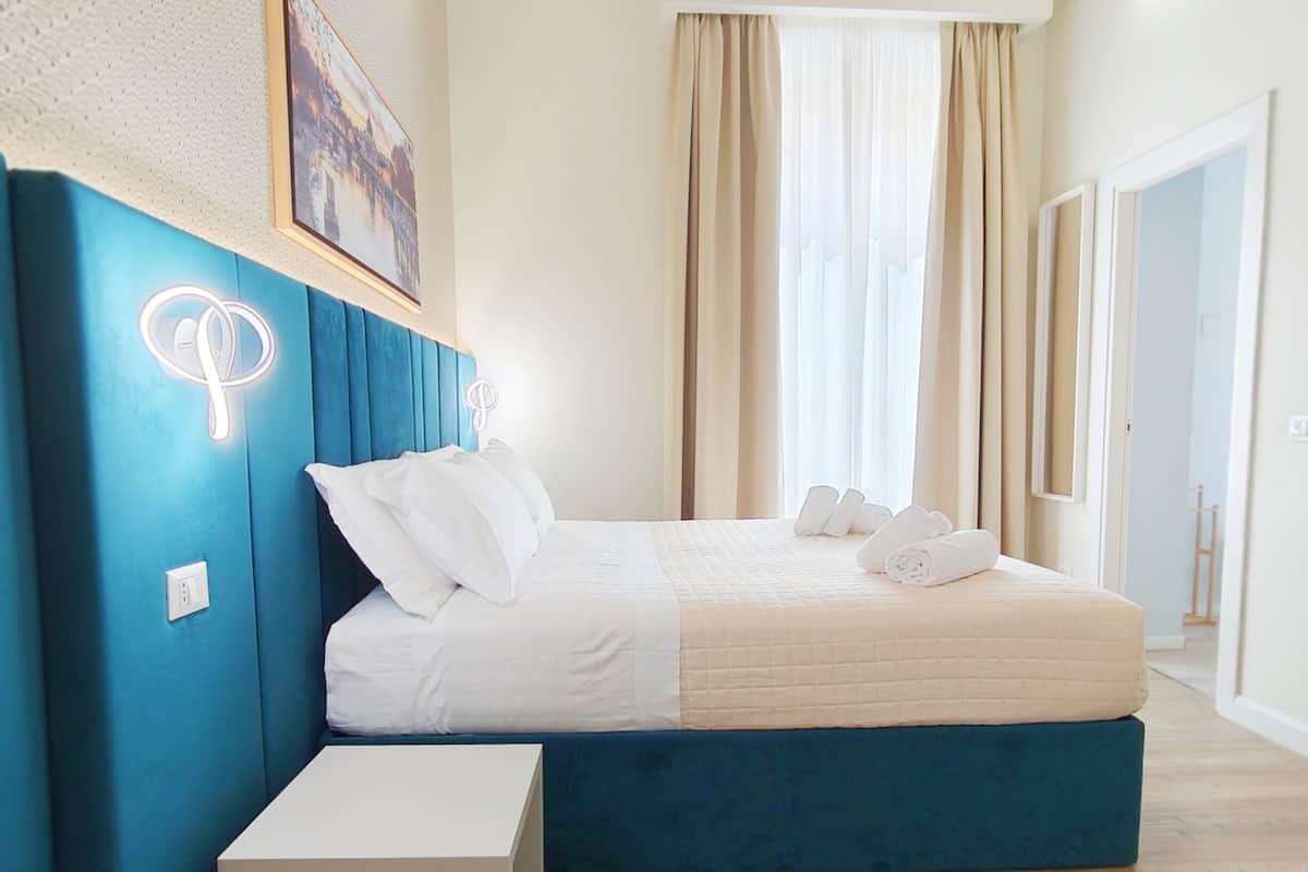 A bedroom with a bed, a bedside table, a headboard, a window with curtains, and a wall-mounted picture.