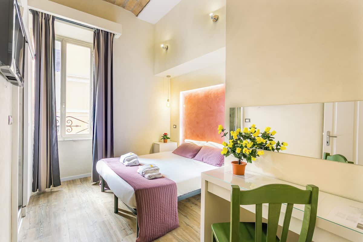 A hotel room with a bed, a desk, a chair, a window with curtains, a potted plant, and a mirror.