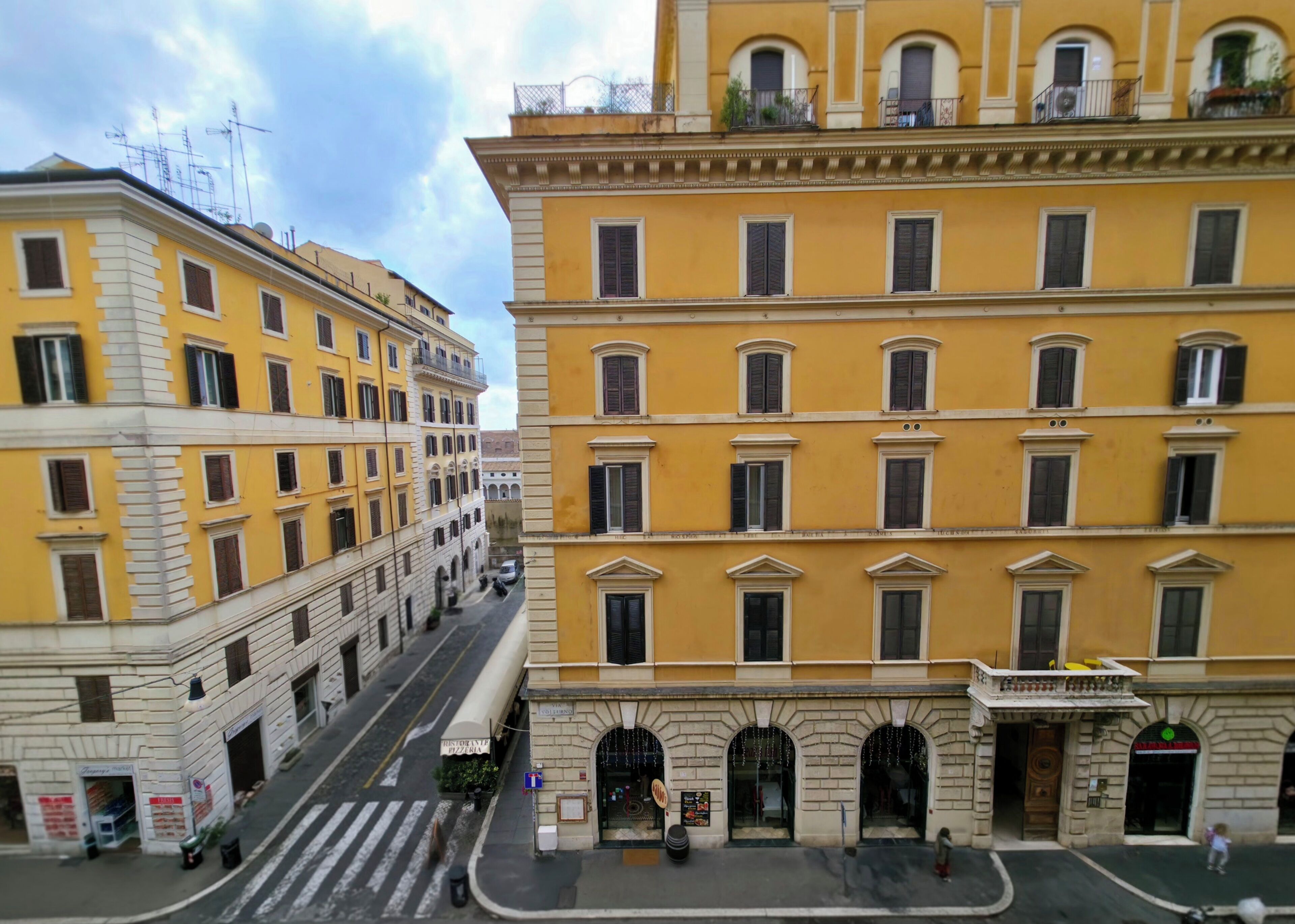 A street view of a yellow building with multiple windows and balconies, a crosswalk, and a few pedestrians.
