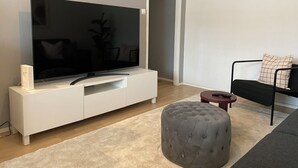 Een moderne woonkamer met een flatscreen televisie op een witte tv-kast, een grijze poef, een zwarte bank met een kussen met dessin, en een kleine ronde tafel.