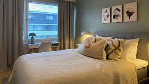 Een slaapkamer met een bed, een bureau, een stoel en een raam met gordijnen. Er hangen drie afbeeldingen in lijsten aan de muur.