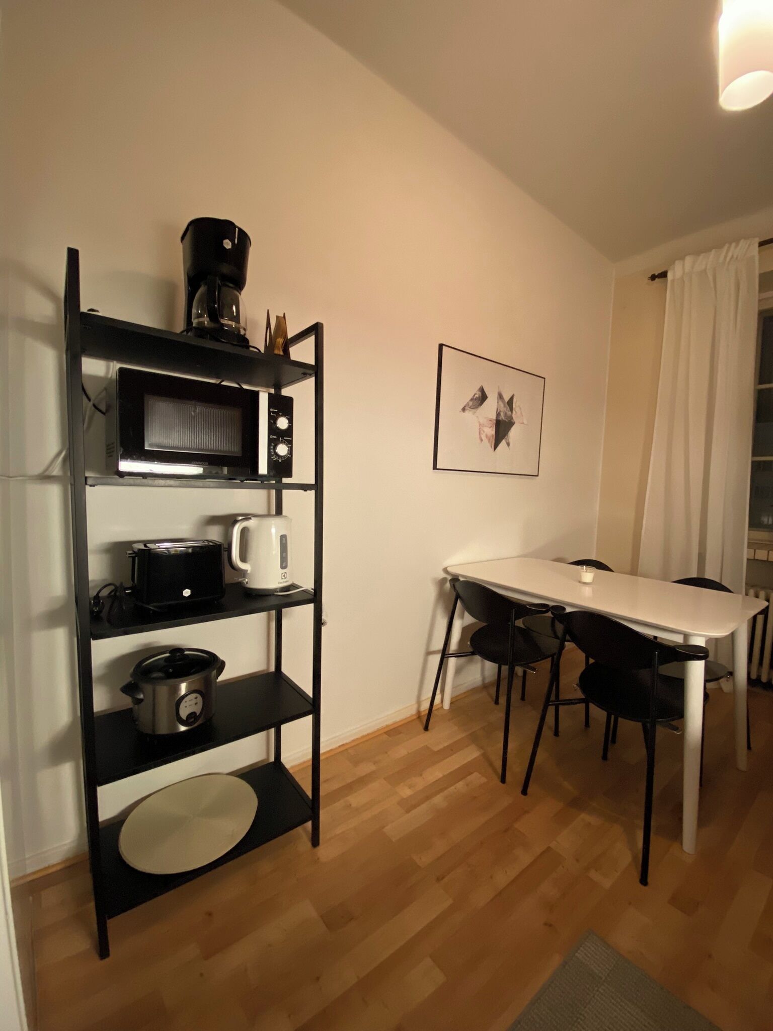 A compact kitchen with a microwave, toaster, and slow cooker on a black shelf unit. A dining table with chairs is adjacent to the kitchen, and a framed picture hangs on the wall.