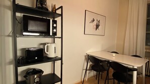 A compact kitchen with a microwave, toaster, and slow cooker on a black shelf unit. A dining table with chairs is adjacent to the kitchen, and a framed picture hangs on the wall. - Homely 1BR in Lively Area with Strategic Location (Helsinki)