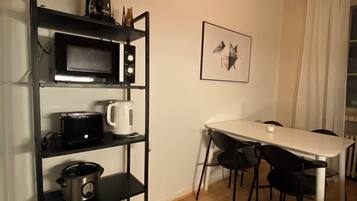 A compact kitchen with a microwave, toaster, and slow cooker on a black shelf unit. A dining table with chairs is adjacent to the kitchen, and a framed picture hangs on the wall.
