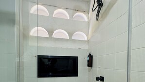 Een douche met een glazen deur, witte betegelde wanden, een vastgeschroefde haardroger en een klein rechthoekig spiegel.