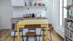 A modern kitchen with wooden flooring, a yellow table, and bar stools. There are plants on shelves, a bookcase, and a window with natural light. - Stylish and modern Studio in Pigalle (Paris)