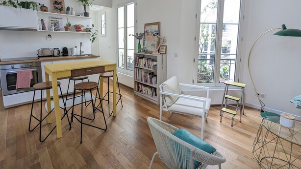A bright kitchen with wooden flooring, a yellow table, and a white chair. There are shelves with plants and books, and a window with a view of the outside. - Stylish and modern Studio in Pigalle (Paris)