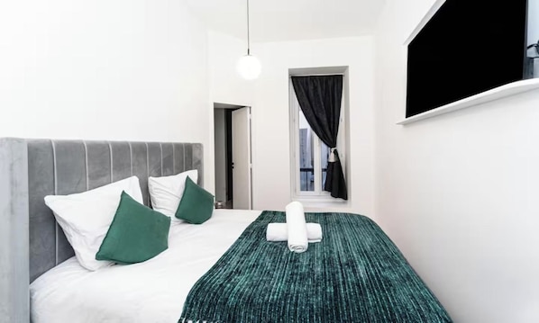 A hotel room with a bed, pillows, a blanket, a window with curtains, and a hanging light fixture.