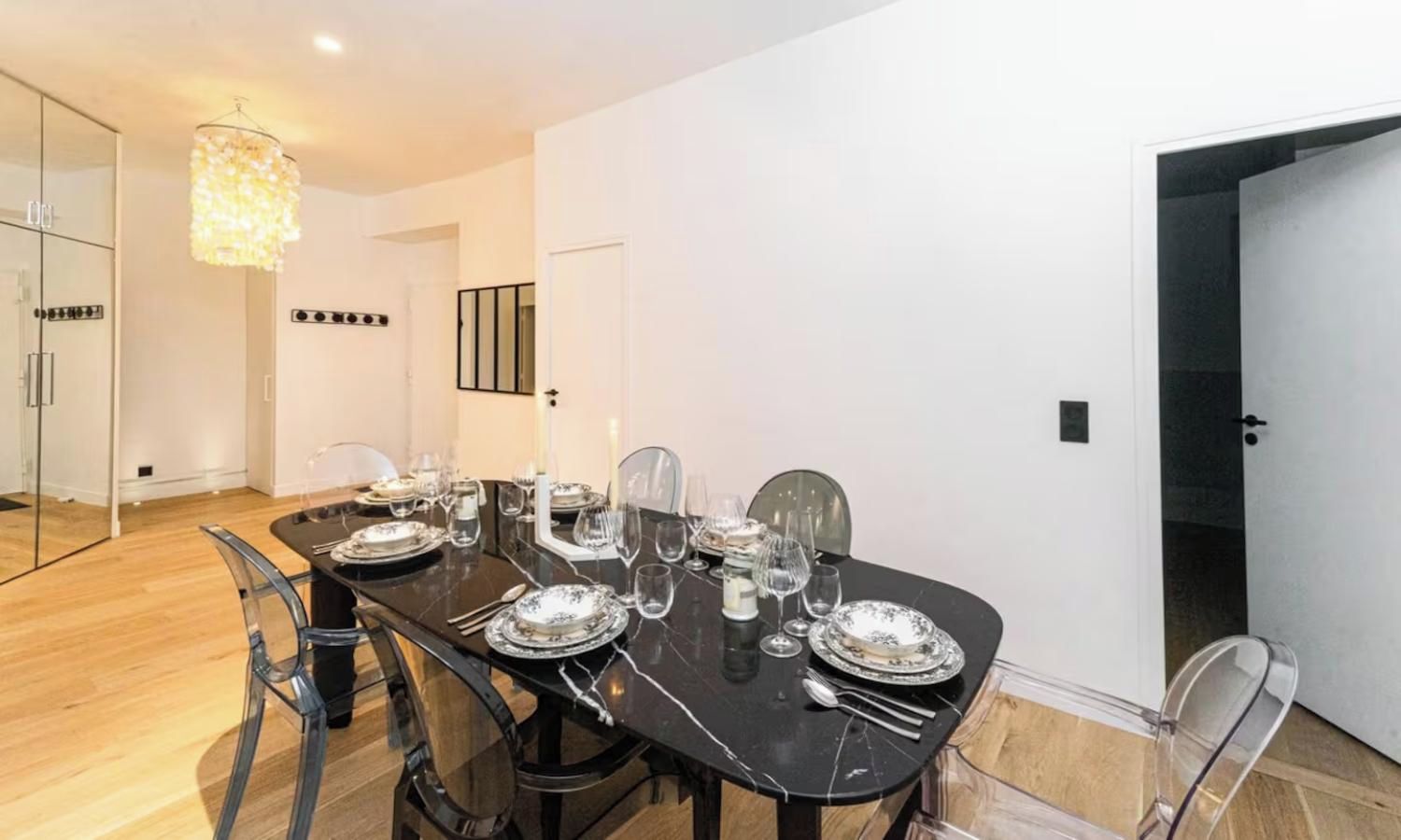 Un comedor con una mesa larga preparada para cuatro personas, sillas traslúcidas y una lámpara de techo moderna.