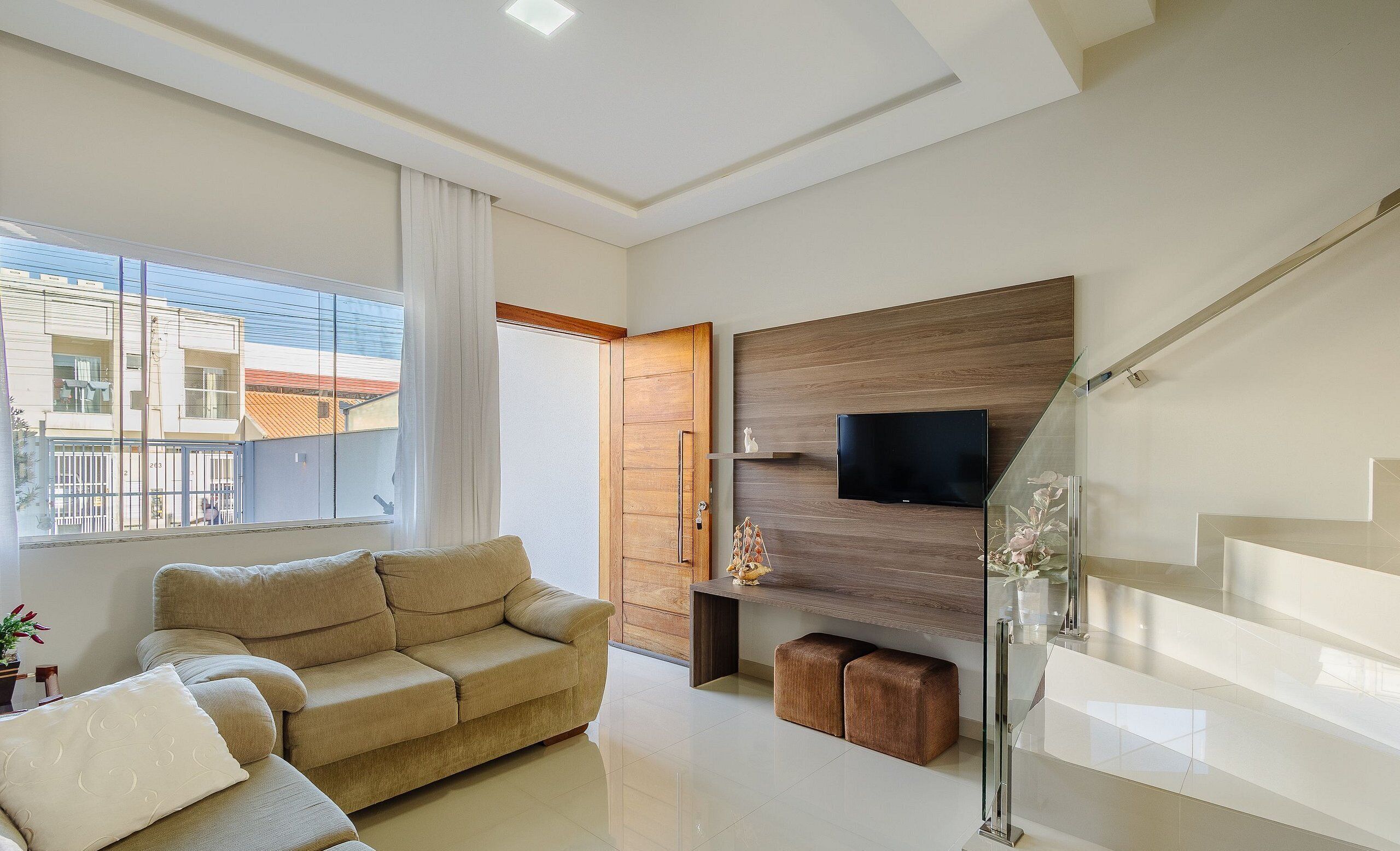 A modern living room with a sofa, a flat-screen TV mounted on a wooden panel, and a glass staircase.