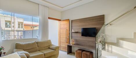A modern living room with a sofa, a flat-screen TV mounted on a wooden panel, and a glass staircase.