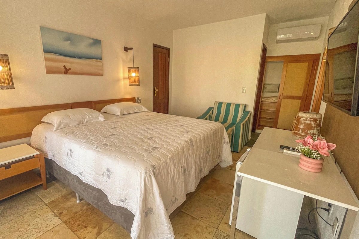 A bedroom with a bed, bedside tables, a desk, a chair, a painting, and an air conditioning unit.