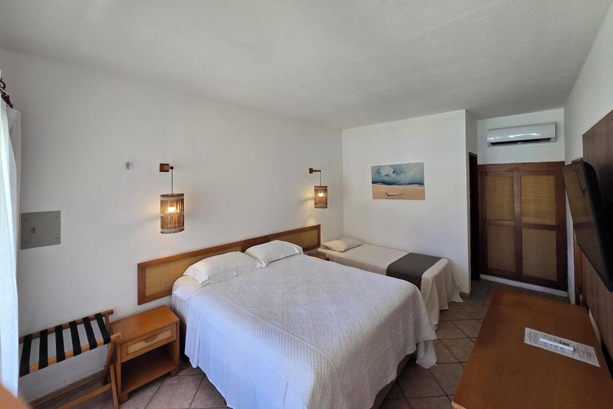 A hotel room with two beds, a wooden desk, a small table, a wall-mounted air conditioner, and a painting.