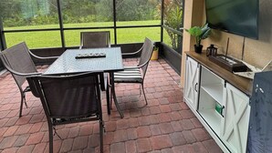 A covered patio area with a table and chairs, a television mounted on the wall, and a view of the garden through large windows.