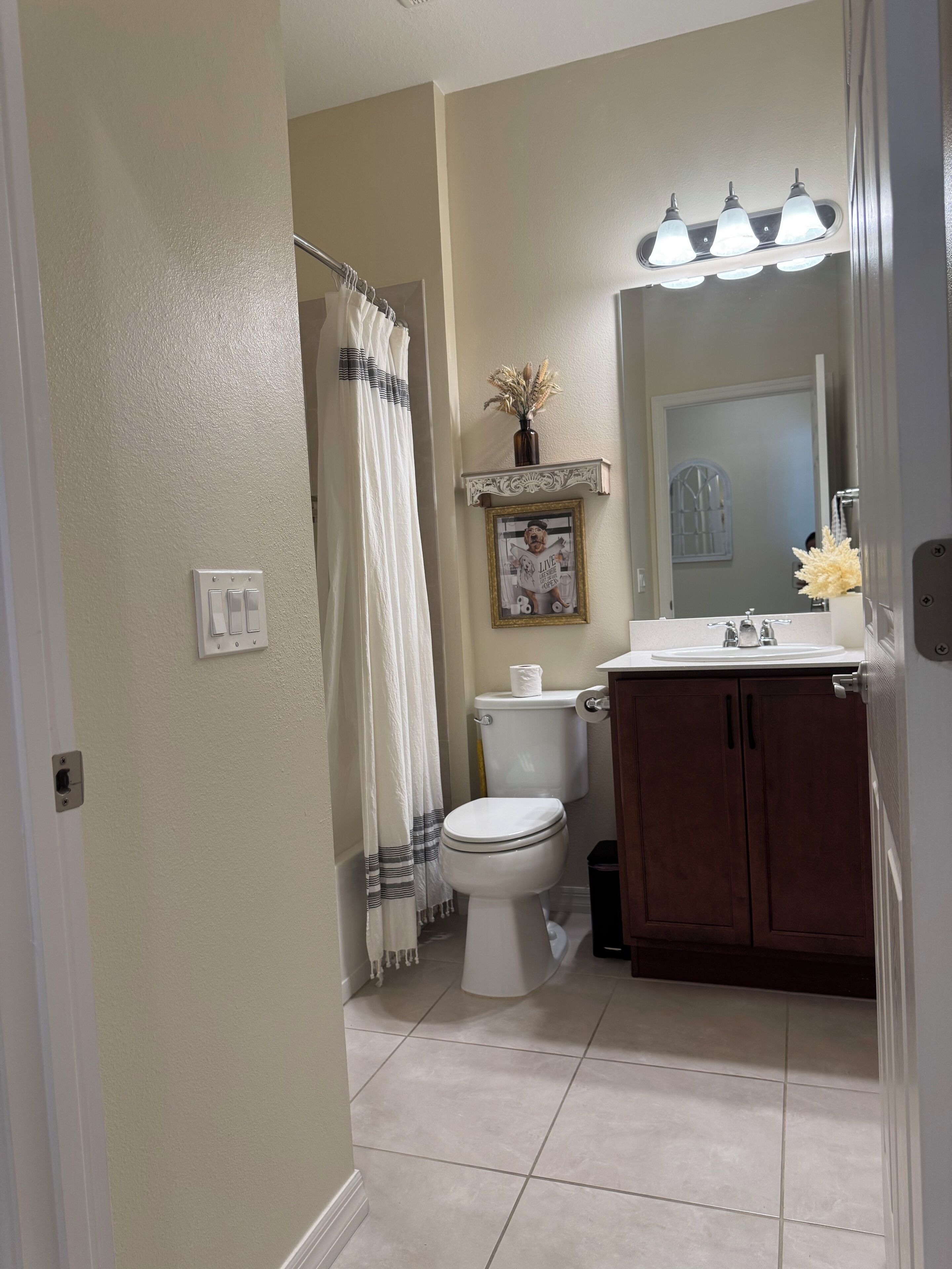Une salle de bain avec des toilettes, un lavabo et un miroir. On y trouve un rideau de douche, un tableau encadré au mur et une petite étagère avec un vase.