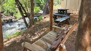 A rustic outdoor seating area with a swing chair and picnic table, overlooking a flowing stream and surrounded by trees.