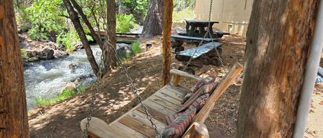 A wooden swing set overlooking a river, with picnic tables and a building in the background.