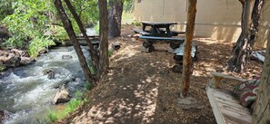 A picnic area with a table and benches near a stream, surrounded by trees and a building in the background.