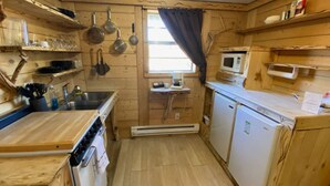 A compact kitchen with wooden walls, a microwave, a small refrigerator, and a window with a curtain.