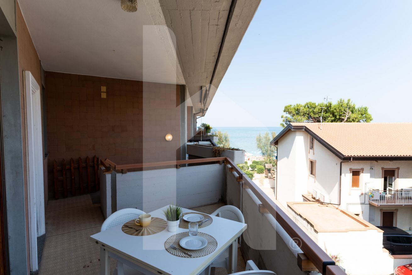 A balcony with a table set for two, overlooking a coastal area with buildings and trees.