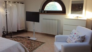Une chambre à coucher avec un lit, un téléviseur, une chaise blanche, un radiateur et une fenêtre donnant sur des arbres.